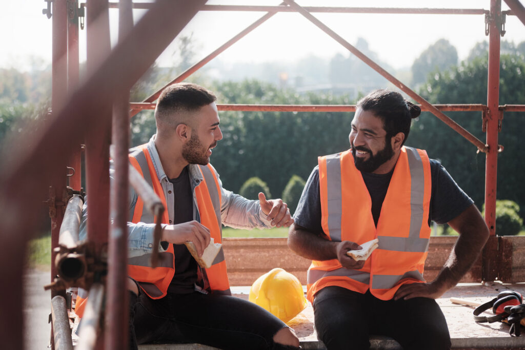 Two Male Construction Workers in orange safety vests eating sandwiches